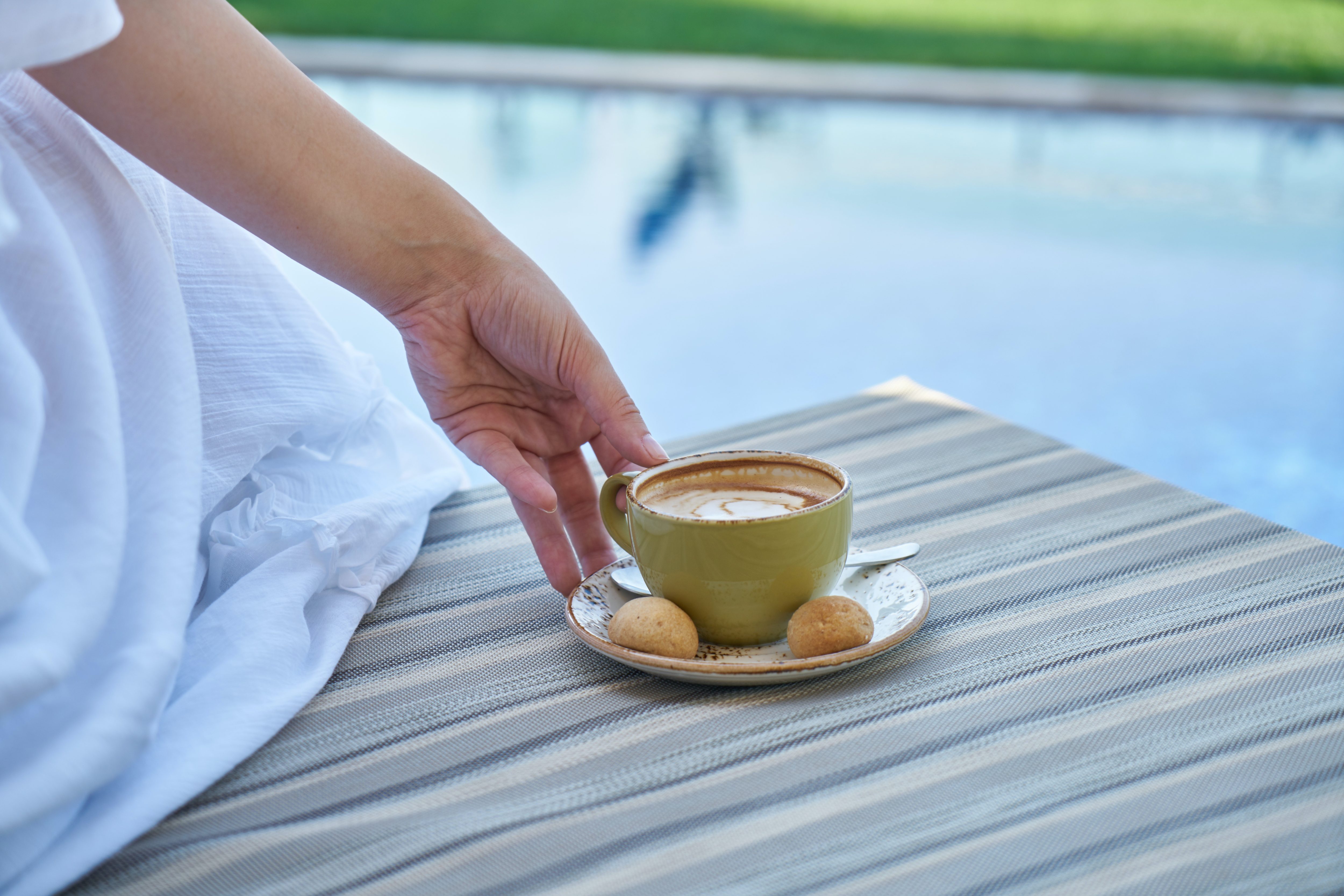 En hand sträcks fram mot en kopp kaffe som står på kanten av en solstol vid en pool.